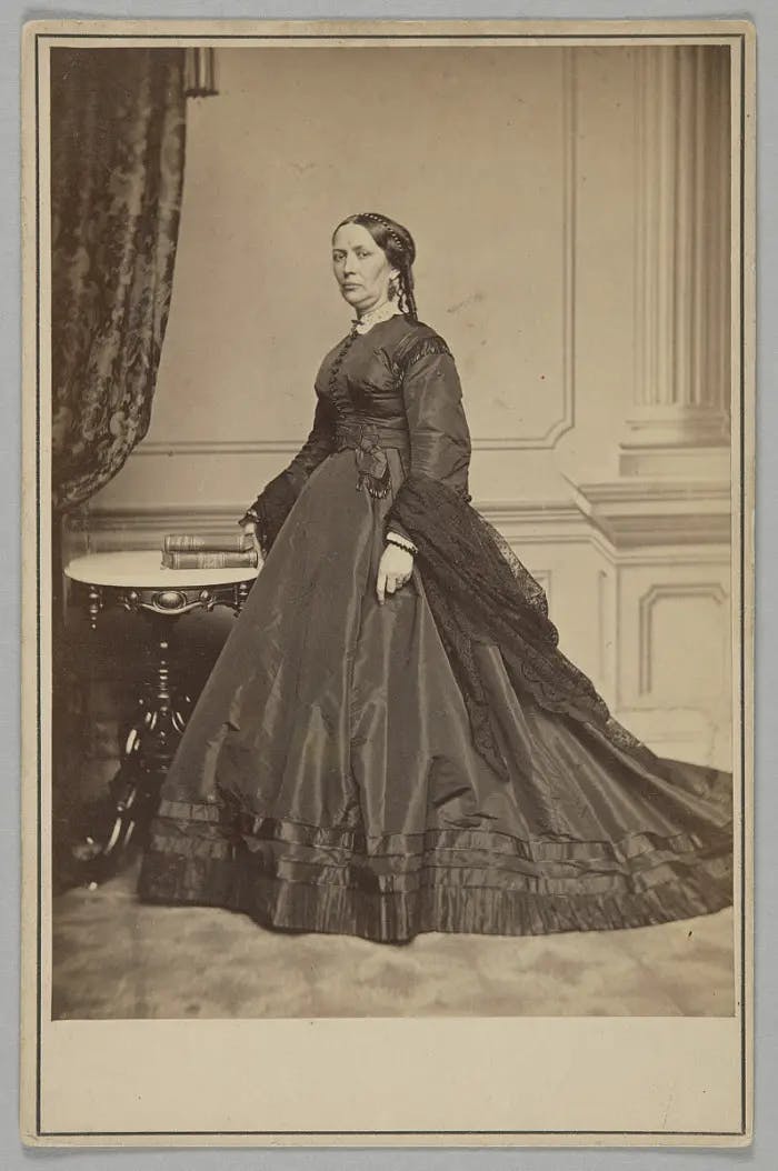 Black and white full body image of MJH Welles wearing a floor-length gown and standing in front of a table in an elegant room.