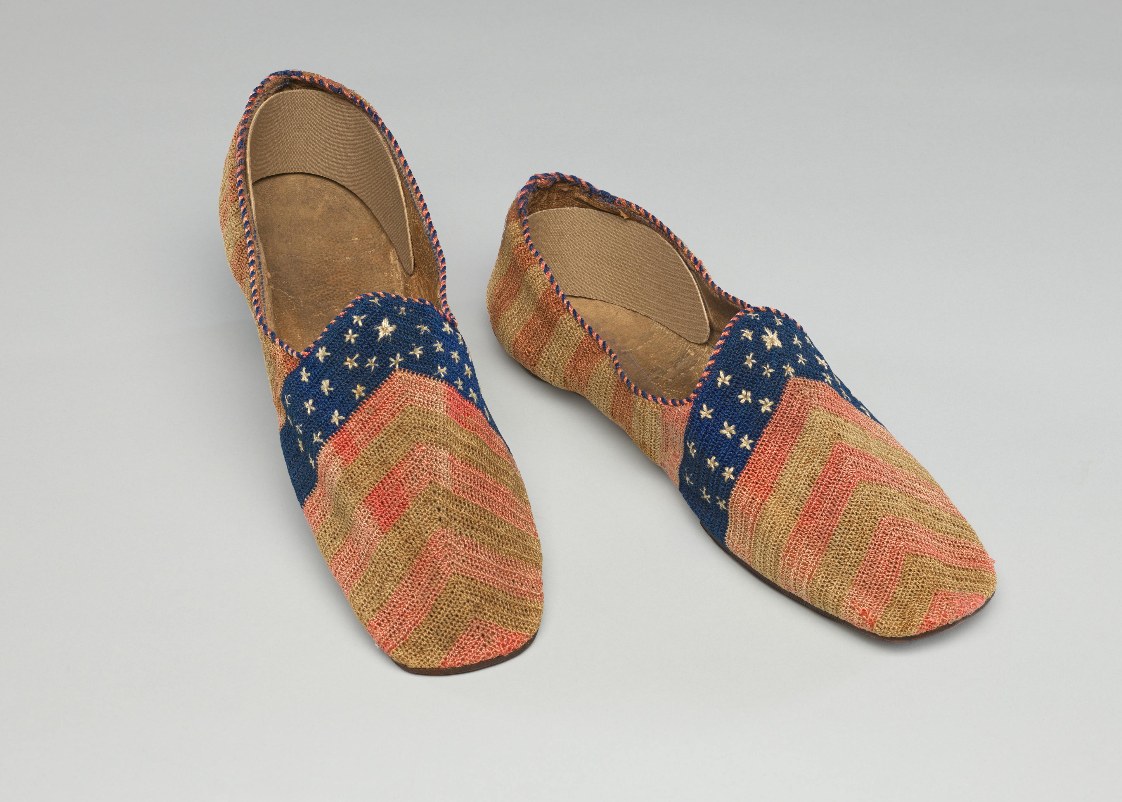 A pair of men's slippers in a stars and stripes design with a flat, leather sole and a squarish toe. The slippers are crocheted in stripes of red and white from the toe to the heel and across the top of the vamp is a blue band embroidered with scattered white stars.