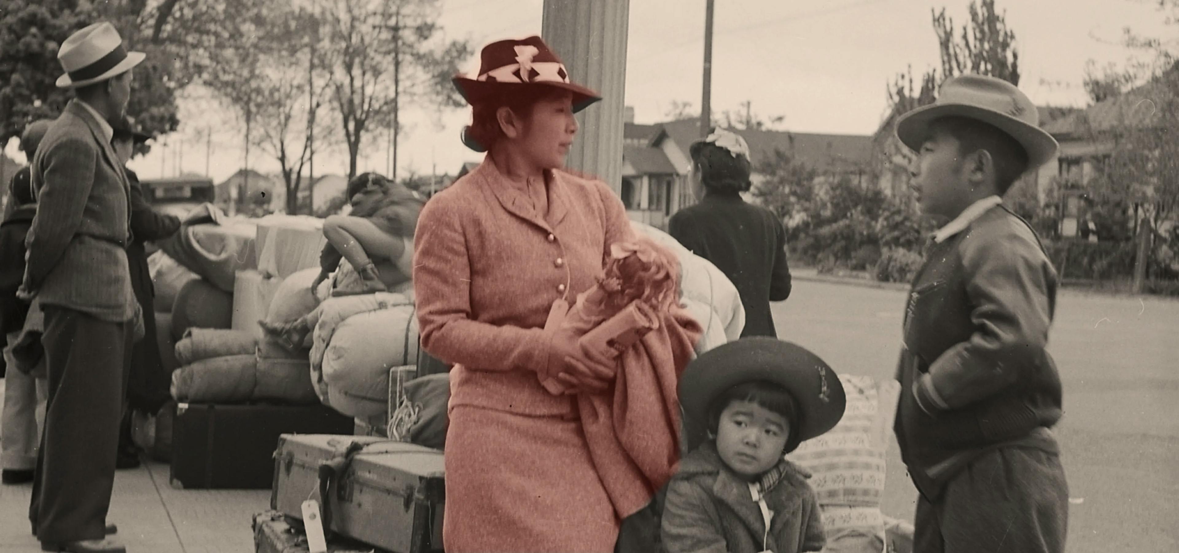 Hisako Hibi and her young daughter stand in front of a pile of luggage, with other people in the background. Hisako is holding her daughter's doll and speaking with a young man. She is in color for emphasis, but the rest of the image is black and white.